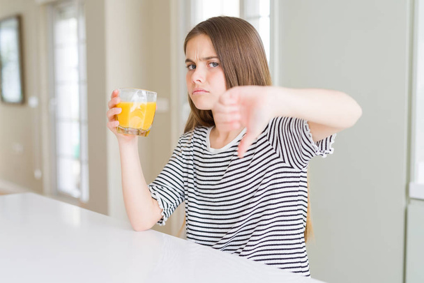 Beautiful young girl kid drinking a glass of fresh orange juice with angry face, negative sign showing dislike with thumbs down, rejection concept - Foto, Imagen