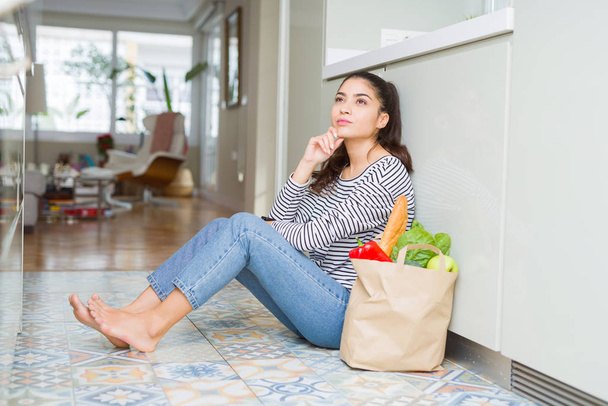 Young woman sitting on the kitchen floor with a paper bag full of fresh groceries with hand on chin thinking about question, pensive expression. Smiling with thoughtful face. Doubt concept. - Photo, Image