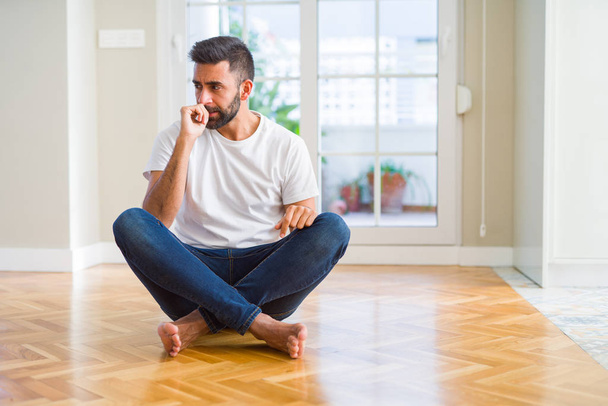 Knappe Spaanse man dragen casual t-shirt zittend op de vloer thuis op zoek gestrest en nerveus met handen op de mond bijten nagels. Angst probleem. - Foto, afbeelding