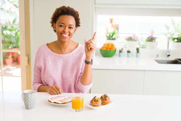 Genç Afro-Amerikalı kadın sabah kahvaltısını evde yiyor. Yüzünde kocaman bir gülümsemeyle, eli ve parmağı kameraya bakıyor.. - Fotoğraf, Görsel