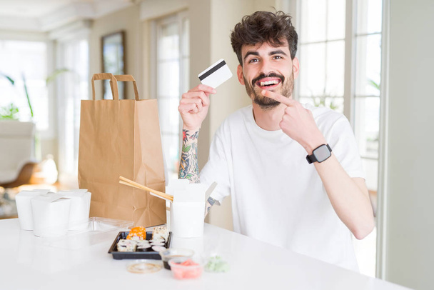 Young man eating asian sushi from home delivery using credit card as payment very happy pointing with hand and finger - Photo, image