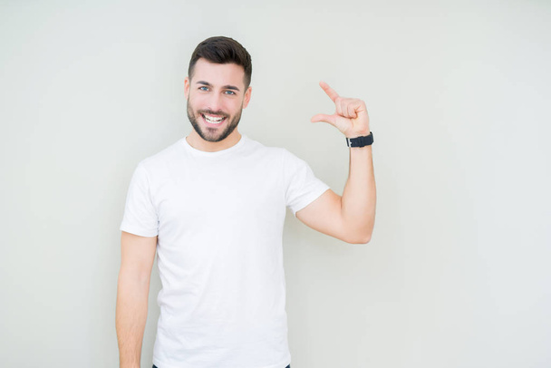 Mladý pohledný muž nosí ležérní bílé tričko usmívá izolovaná pozadí a jistý ukázal rukou dělá znamení velikost prsty při pohledu a fotoaparát. Opatření koncepce. - Fotografie, Obrázek