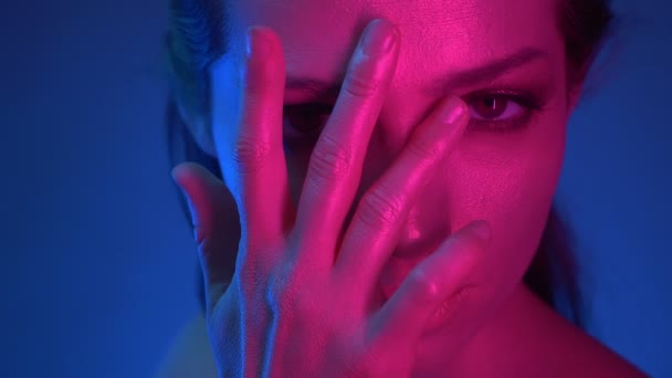 Close-up shoot van model met glitter make-up in blauw en roze neonlichten bedekt haar gezicht met vingers kijken naar camera scherp. - Video