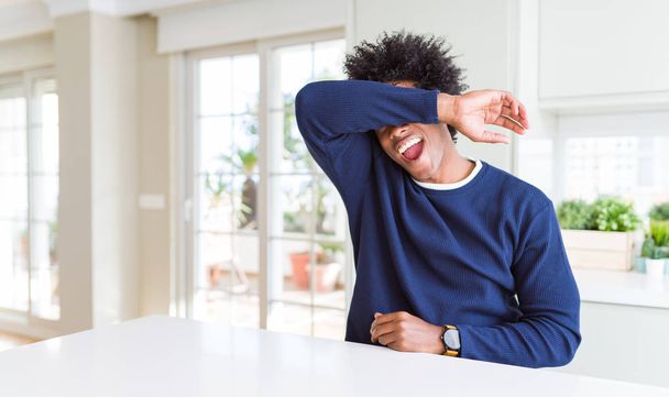 若いアフリカ系アメリカ人の男性は、陽気で面白い腕で目を覆う家に座ってカジュアルなセーターを着ています。ブラインドコンセプト. - 写真・画像