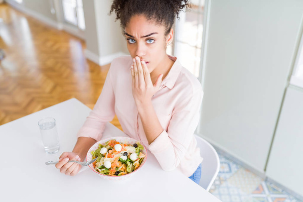 Jonge african american vrouw eten gezonde pasta salade cover mond met de hand geschokt met schaamte voor fout, uitdrukking van angst, bang in stilte, geheime concept - Foto, afbeelding