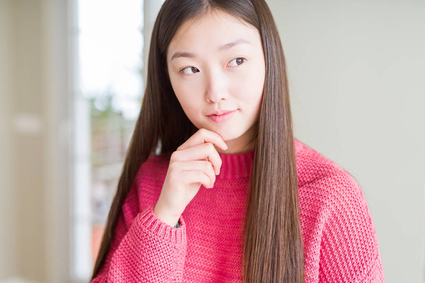 Beautiful Asian woman wearing pink sweater with hand on chin thinking about question, pensive expression. Smiling with thoughtful face. Doubt concept. - Photo, image