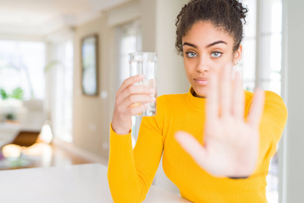 Young african american woman drinking a glass of fresh water with open hand doing stop sign with serious and confident expression, defense gesture - Foto, Imagen