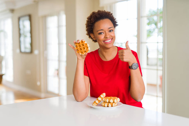 若いアフリカ系アメリカ人女性が大きな笑顔で幸せな甘いワッフルを食べてOKサイン、指で親指アップ、優れた兆候 - 写真・画像