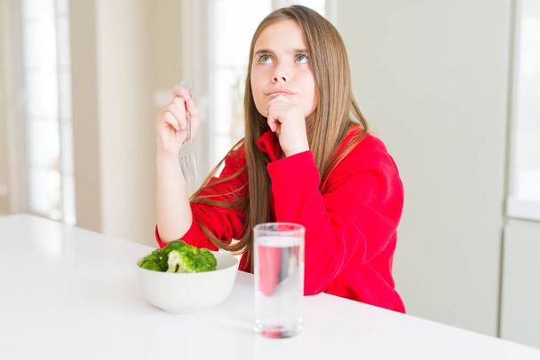 Beautiful young girl kid eating fresh broccoli and drinking water serious face thinking about question, very confused idea - Photo, Image
