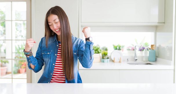 Beautiful Asian woman wearing denim jacket on white table excited for success with arms raised celebrating victory smiling. Winner concept. - Фото, изображение