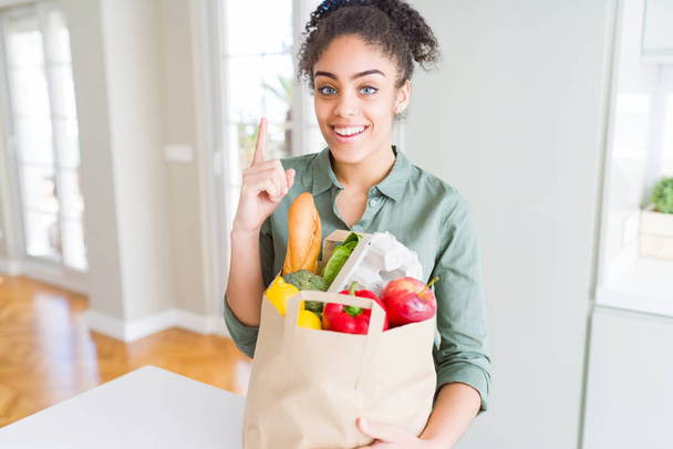 Молодая африканская американка с бумажным пакетом продуктов из супермаркета удивлена идеей или вопросом, указывая пальцем со счастливым лицом, номер один
 - Фото, изображение