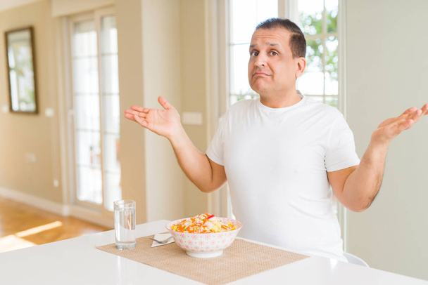 Homem de meia idade comendo arroz em casa sem noção e expressão confusa com braços e mãos levantadas. Conceito de dúvida
. - Foto, Imagem