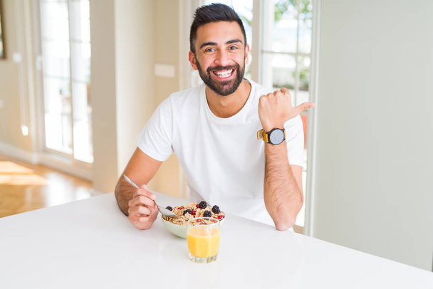 Handsome hispanic man eating healthy breakfast in the morning at home pointing and showing with thumb up to the side with happy face smiling - Фото, изображение