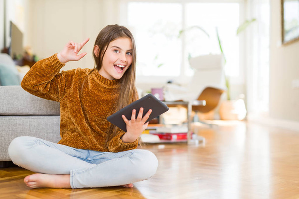 Piękna młoda dziewczyna dziecko za pomocą cyfrowego panelu dotykowego tabletu, siedząc na podłodze zaskoczony z pomysłu lub pytanie palcem wskazującym z szczęśliwy twarz, numer jeden - Zdjęcie, obraz