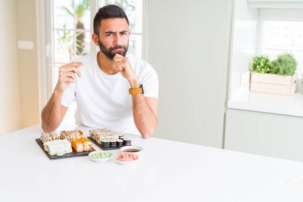 Hombre hispano guapo comiendo sushi asiático usando palillos cara seria pensando en la pregunta, idea muy confusa
 - Foto, imagen