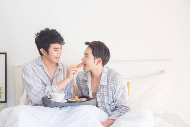 Joven pareja gay vistiendo pijamas sentado en la cama y desayunando
 - Foto, imagen