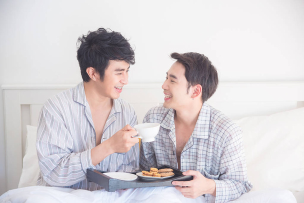 Νεαρό γκέι ζευγάρι που φοράει πιτζάμες κάθεται στο κρεβάτι και τρώει πρωινό - Φωτογραφία, εικόνα
