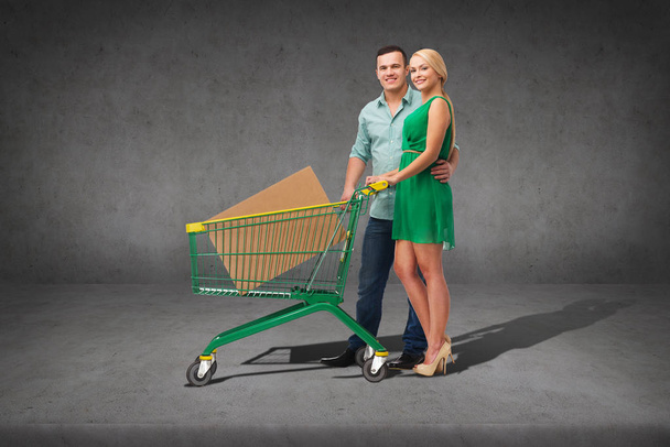 happiness,electronics, shopping and couple concept - smiling couple with shopping cart and big cardboard box in it - Foto, Imagem
