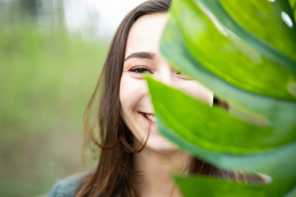 Beautiful natural young woman closeup behind big monstera leaf with green background in the woods. Beauty, health and nature concept. - Fotografie, Obrázek