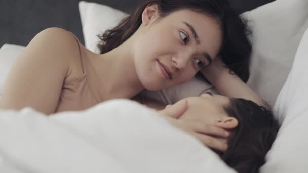 Lesbian couple hugging and smiling while lying together in bed at home. Young lesbians kisses and hugs after wake up - Záběry, video