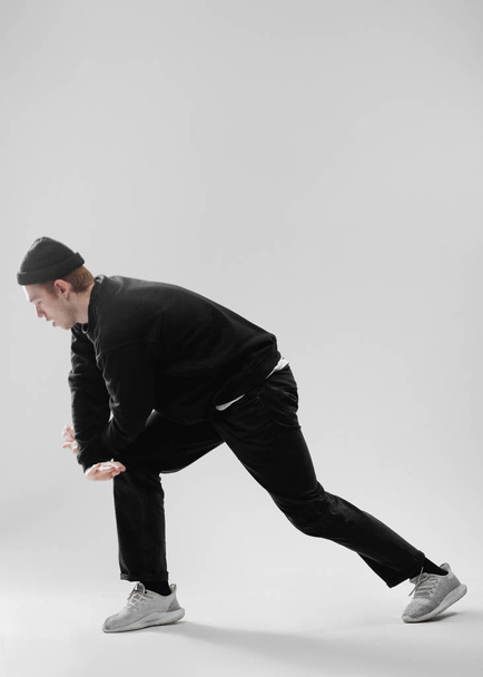 Guy dancer dressed in black jeans, sweatshirt, hat and gray sneakers is dancing in the studio on the white background - Fotó, kép