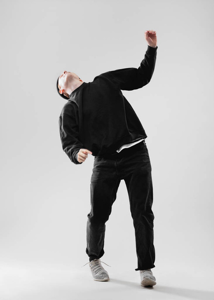 Dancer dressed in black jeans, sweatshirt, hat and gray sneakers is dancing  in the studio on the white background - Фото, изображение