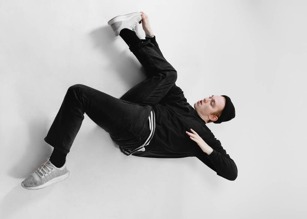 Freestyle dancer dressed in black jeans, sweatshirt, hat and gray sneakers is dancing lying on the floor in the studio on the white background - Photo, Image