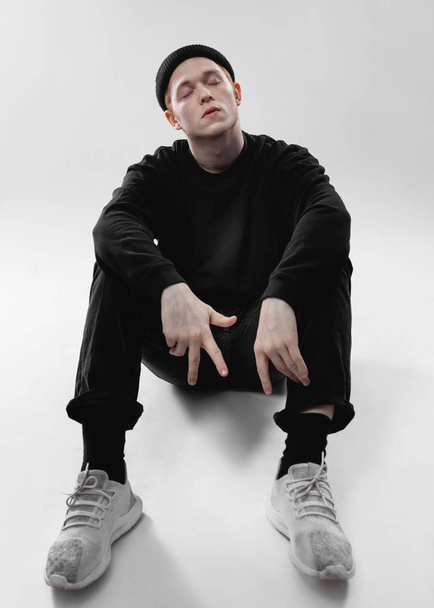Guy dressed in black jeans, sweatshirt, hat and gray sneakers is sitting on the floor in the studio on the white background - Foto, Imagem