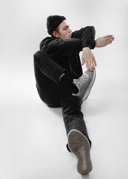 Freestyle dancer dressed in black jeans, sweatshirt, hat and gray sneakers is dancing sitting on the floor in the studio on the white background - Fotografie, Obrázek