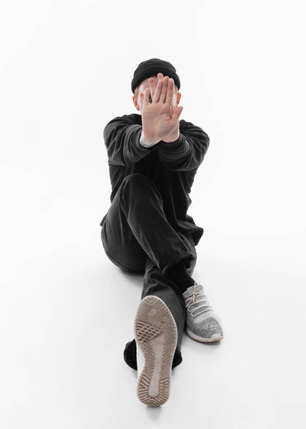 Freestyle dancer dressed in black jeans, sweatshirt, hat and gray sneakers is dancing sitting on the floor in the studio on the white background - Φωτογραφία, εικόνα