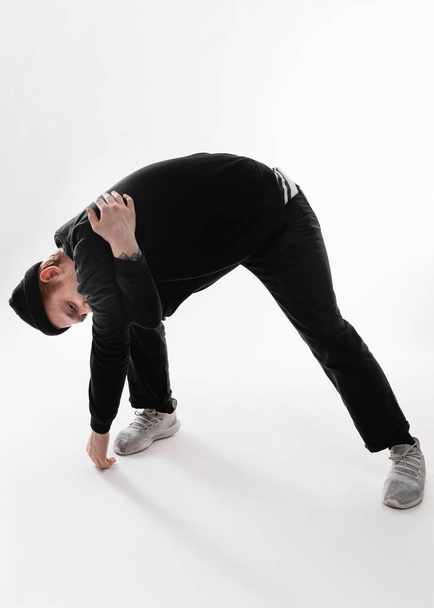 Guy dressed in black jeans, sweatshirt, hat and gray sneakers stands leaning forward in the studio on the white background - Photo, image