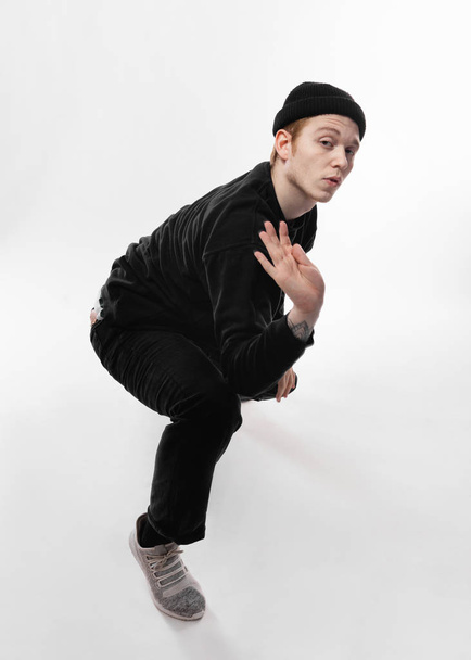 Young freestyle dancer dressed in black jeans, sweatshirt, hat and gray sneakers is dancing in the studio on the white background - Photo, Image