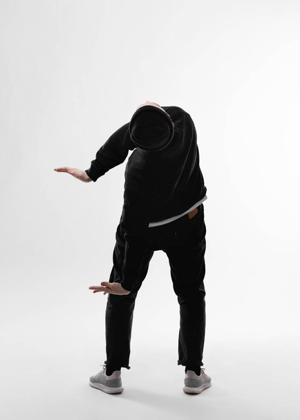 Young freestyle dancer dressed in black jeans, sweatshirt, hat and gray sneakers is dancing  closing his face with his hand in the studio on the white background - Foto, Imagem