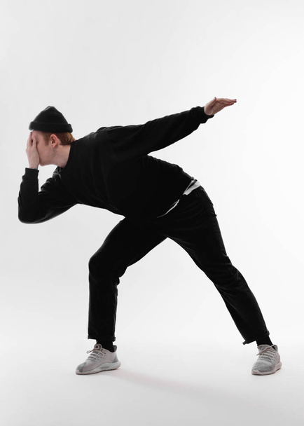 黒いジーンズ、スウェットシャツ、帽子、グレーのスニーカーを身に着けている若いフリースタイルのダンサーは、白い背景のスタジオで手で顔を閉じて踊っています - 写真・画像