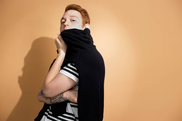 Stylish red-haired guy in a striped shirt with tattoo on his hand is posing with black sweater on his shoulders on the beige background in the studio - 写真・画像