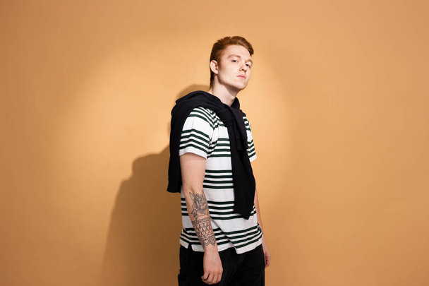 Stylish red-haired guy in a striped shirt with tattoo on his hand is posing with black sweater on his shoulders on the beige background in the studio - Foto, Bild