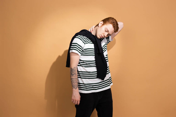 Stylish red-haired guy in a striped shirt with tattoo on his hand is posing with black sweater on his shoulders on the beige background in the studio - Foto, imagen