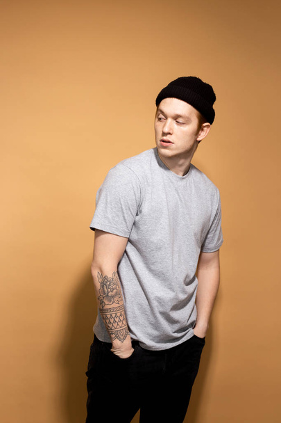 Stylish guy dressed in a gray shirt, black jeans and black hat with tattoo on his hand is posing on the beige background in the studio - Fotografie, Obrázek