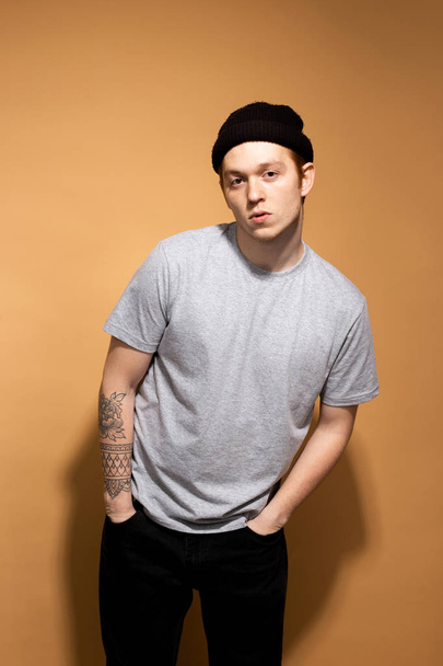Young stylish guy dressed in a gray shirt, black jeans and black hat with tattoo on his hand is posing on the beige background in the studio - Foto, afbeelding