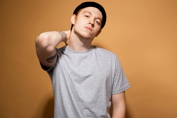 Young stylish guy dressed in a gray shirt and black hat with tattoo on his hand is posing on the beige background in the studio - Photo, image