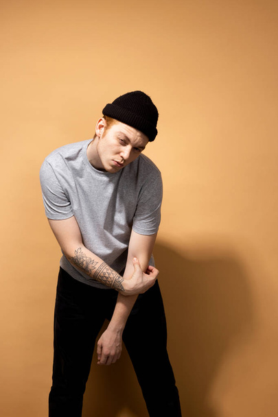 Young stylish guy dressed in a gray shirt, black jeans and black hat with tattoo on his hand is posing on the beige background in the studio - Fotó, kép