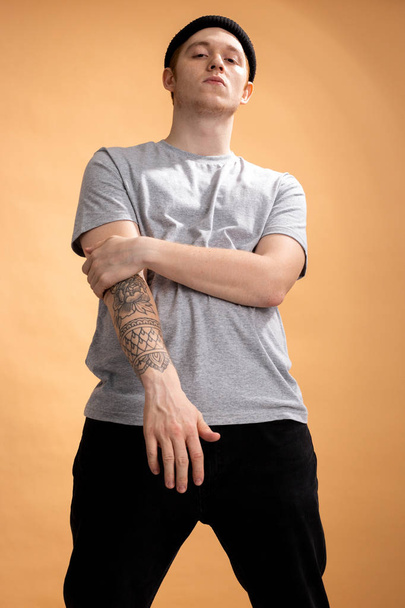 Cocky stylish guy dressed in a gray shirt, black jeans and black hat with tattoo on his hand is standing on the beige background in the studio - Foto, afbeelding