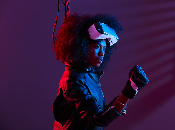 Stylish curly dark haired girl dressed in black leather jacket and gloves is wearing the virtual reality glasses on her head in the dark studio with neon light - Fotoğraf, Görsel