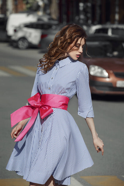 Beautiful stylish brunet girl dressed in striped white and blue dress with a bright pink belt with a bow at the waist is walking in the street on a summer day - Foto, Bild