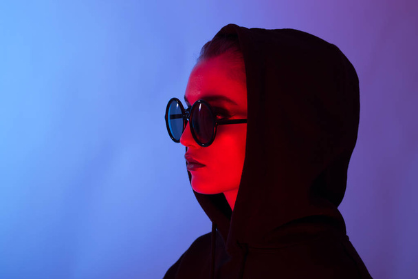 Portrait of fashion young girl in a black sweater with a hood and round sunglasses in red and blue neon light in the studio - Photo, image