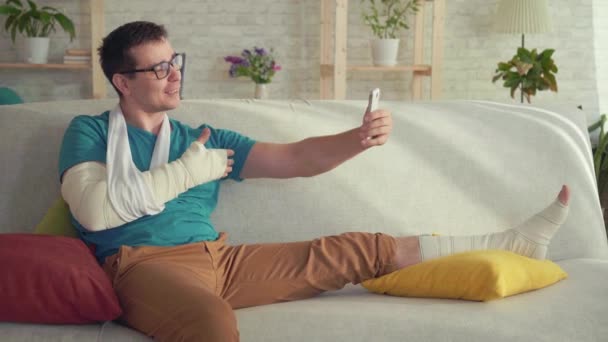 Young man with a broken arm and leg sitting on the couch talking on video call - Footage, Video