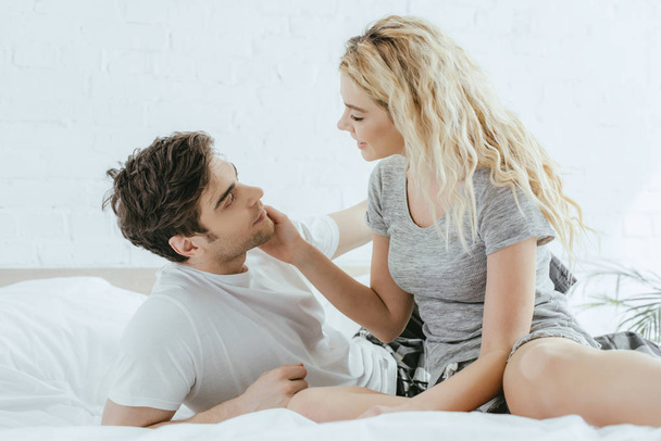 happy blonde woman touching cheek of handsome man lying on bed  - 写真・画像