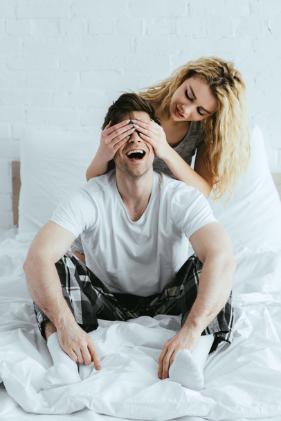 ελκυστική ξανθιά κορίτσι κλείνει τα μάτια του ευτυχισμένο άντρα κάθεται στο κρεβάτι  - Φωτογραφία, εικόνα
