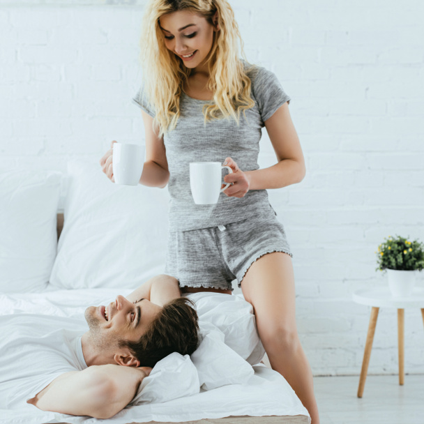 vidám szőke lány gazdaság csésze kávé közelében boldog ember fekve az ágyban  - Fotó, kép