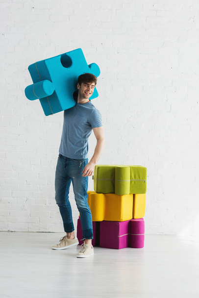 cheerful man standing and holding blue jigsaw piece near white brick wall  - Foto, Imagen